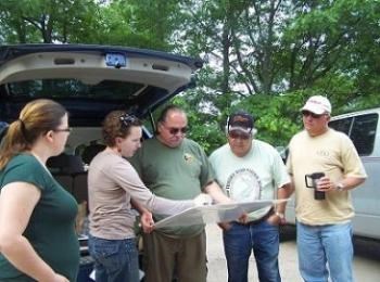 Five people looking at documents near vehicles.