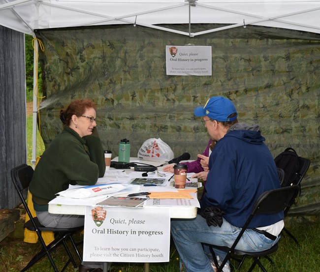 An NPS historian and a male event visitor sit across from each other at a table inside a tent and talk