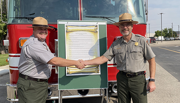 Two men shake hands in front of a fire engine and printed proclamation.