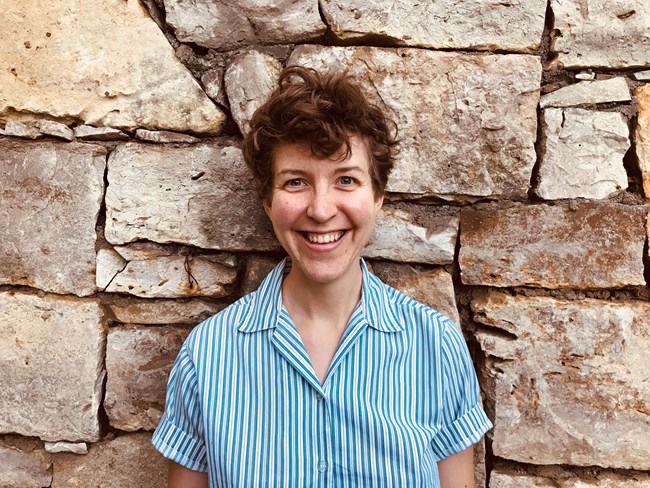 Dr. Sophia Ford A person with brown hair and a blue striped shirt standing in front of a stone wall.