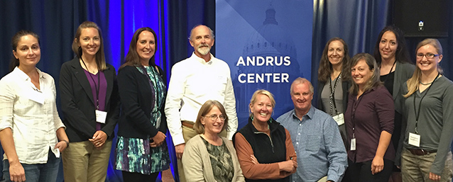 Men and women gathered in front of a blue curtain around an Andrus Center banner.