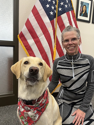 A yellow dog (left) sits and a woman in a gray dress (right) kneels in front of the American flag.