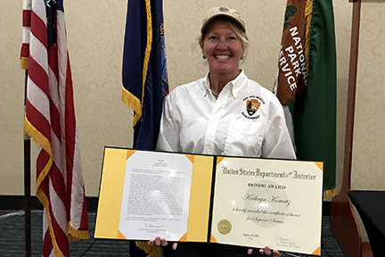 Kathy Komatz stands in front of US, DOI, and NPS flags holding certificate.