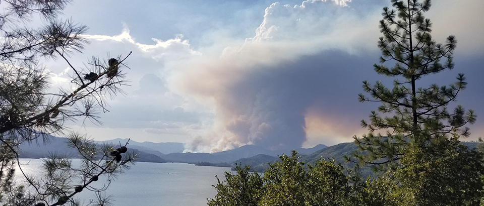 A large plume of smoke rises from a forested slope beyond a lake.