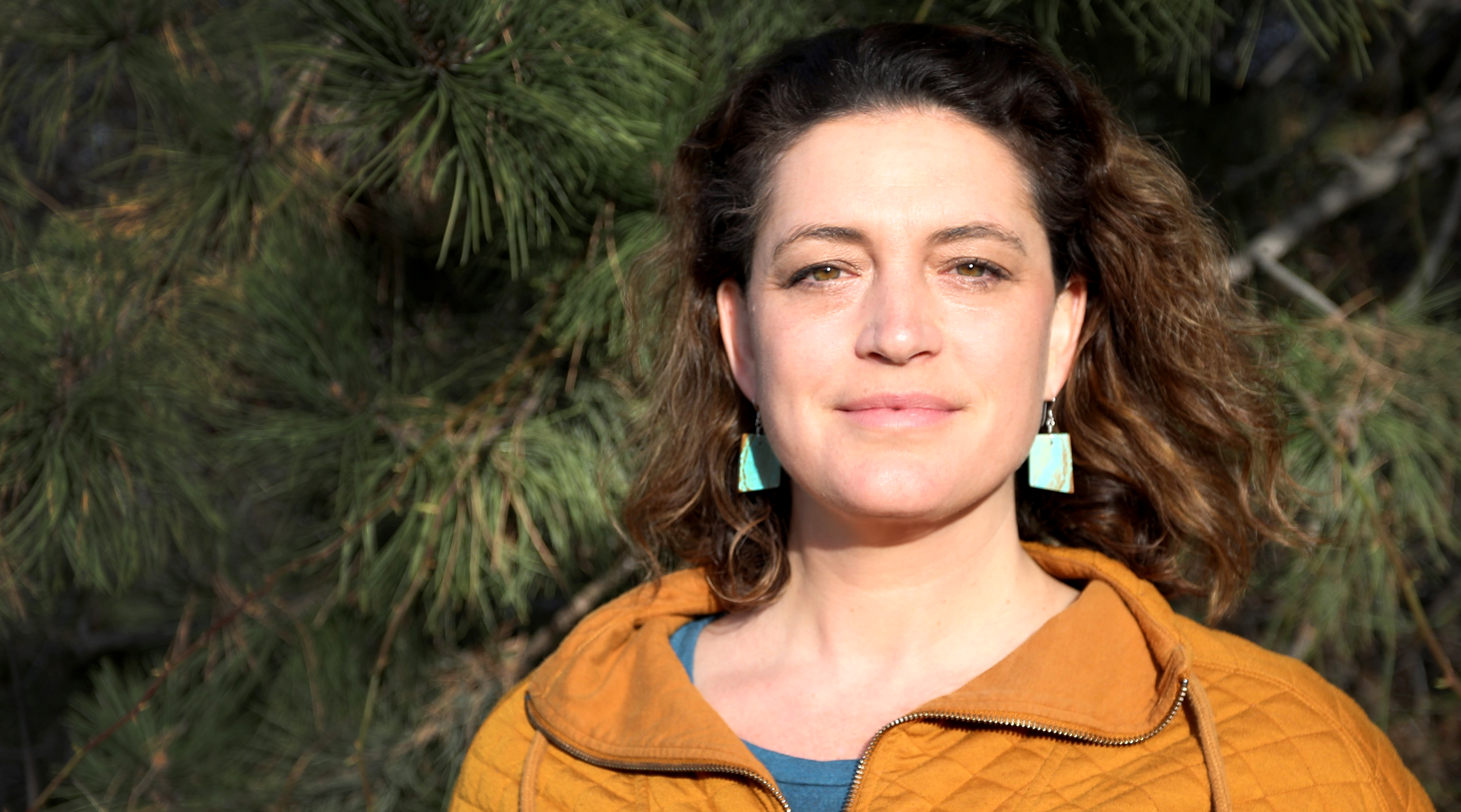 Rhiannon Touchette. A dark-haired young woman with dangling turquoise earrings and hazel eyes stands in front of a dark tree.
