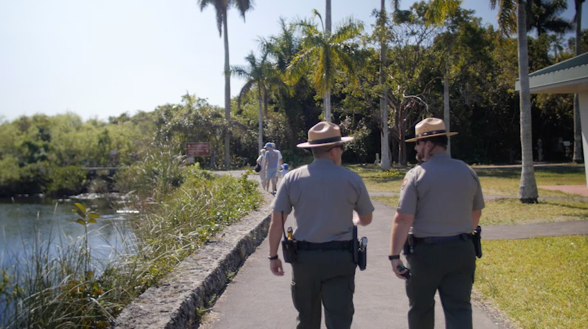 Pasted_image_at_2018-09-04__11_29_AM Two rangers walking down a path next to a body of water with members of public ahead of them in the distance.