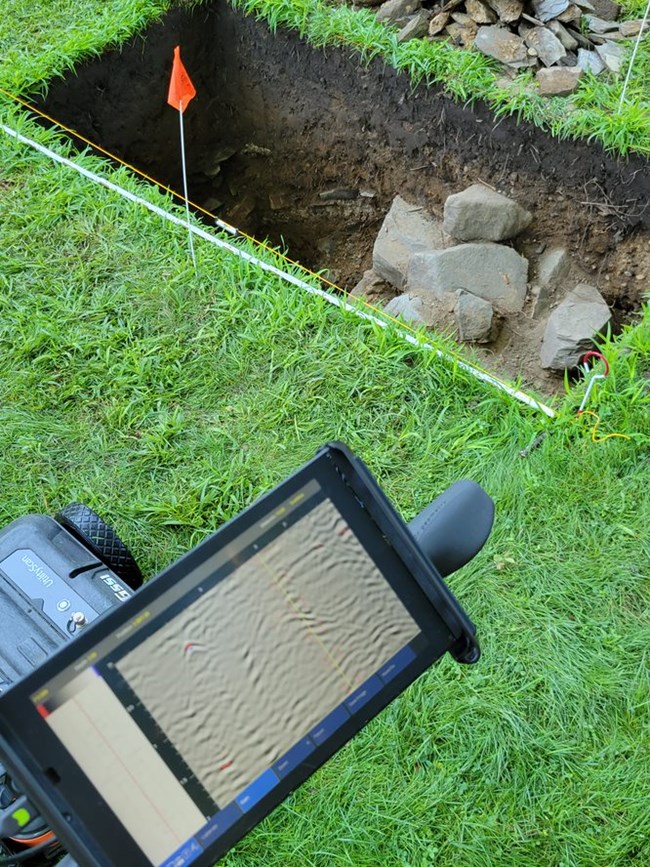 GPR Machine in front of an excavation unit. A GPR machine with a scanner sits next to an open excavation unit with rocks peeking out