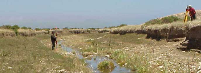A shallow creek flows through a grassy eroded gully. One man stands by the water holding a red and white striped pole. Another man stands on the small bluff above the water with a surveyor's tripod. A shallow creek flows through a grassy eroded gully. One man stands by the water holding a red and white striped pole. Another man stands on the small bluff above the water with a surveyor's tripod.