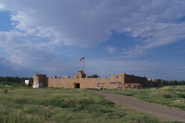 A large stucco building in a green field, with a dirt path leading to it. There is a teepee in the front and a chuckwagon to the side.