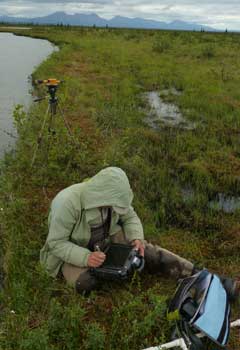 A researcher sits on mossy ground next to a lake with mountains in the far distance over a large area of flat tundra. She enters data into a small screen on her lap. A researcher sits on mossy ground next to a lake with mountains in the far distance over a large area of flat tundra. She enters data into a small screen on her lap.