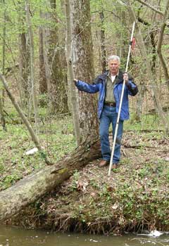 A man in jeans and a raincoat stands in a wooded area along a river holding a tall narrow pole. A man in jeans and a raincoat stands in a wooded area along a river holding a tall narrow pole.
