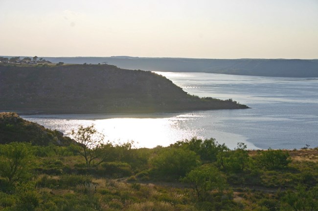 The setting sun reflects off the surface of a lake. A peninsula reaches out into the lake and shrubs are seen in the foreground.