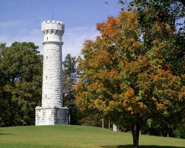 A tall white monument set upon a grassy knoll and flanked by deciduous trees in early autumn.