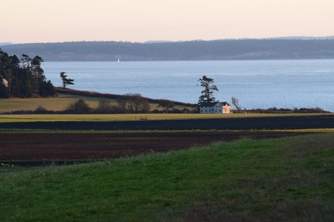 NRCA EBLA An old house sits between rich farmland and the sea. A sailboat can be seen in the distance.