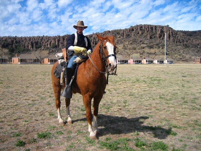 A man wearing a blue uniform sitting on a brown horse with buildings and a rocky hill in the background.