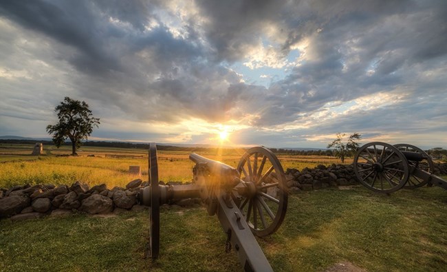 The sun sets over the battlefield. Two cannons sit in front of a stone wall and a monument and a tree are in the left distance.