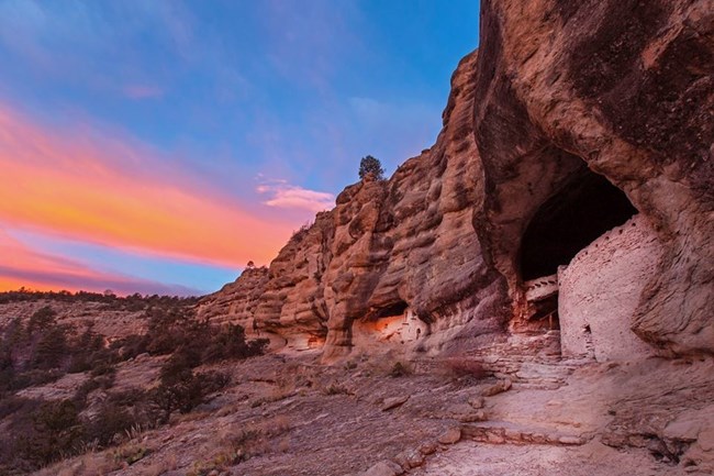 Clay dwellings in the side of a rock cliff with colorful orange sunset-lit clouds in the sky.