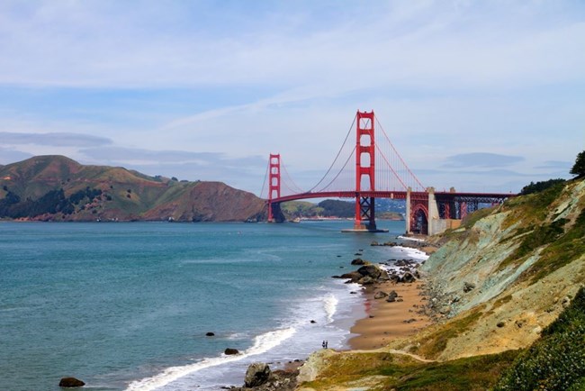 Ocean and rocky coastline surrounded by hills and the red-orange golden gate bridge in the background.