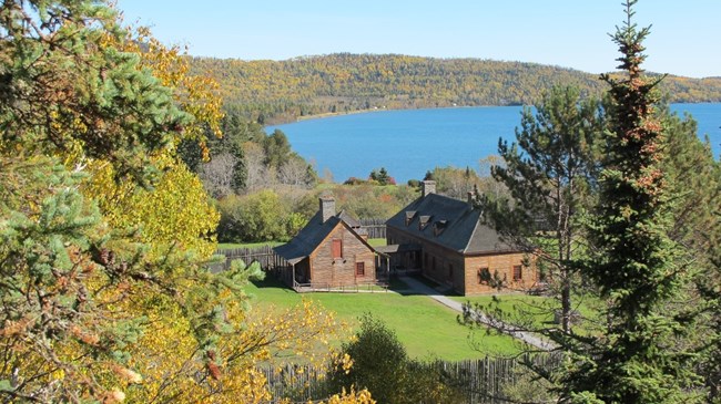 Looking through trees at a wooden depot in a green field, with a blue Lake Superior in the background.