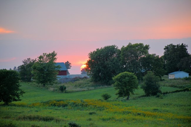 A green hill with trees in front of a red building and the sun setting in the background.