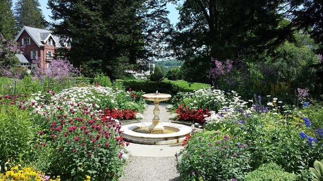 Colorful flowers bloom around a fountain, and a brick mansion is seen in the background.