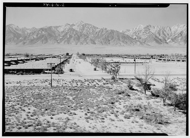 Black and white historical photo of mumerous barrack buildings are lined up in the foreground with mountains in the background.