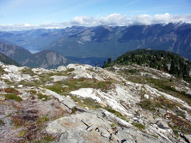 A cloud-topped mountain range and the glimpse of a lake can be seen from a rocky mountain trail.