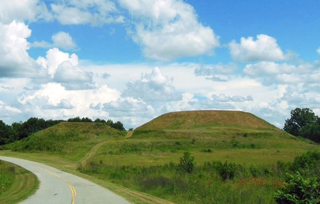 A view of two green mounds from a curved road with a cloudy sky above