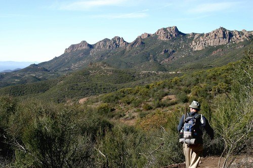 A hiker traverses a long trail through woody shrublands at the base of mountain peaks.