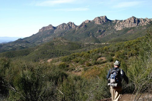NRCA SAMO A hiker traverses a long trail through woody shrublands at the base of mountain peaks.