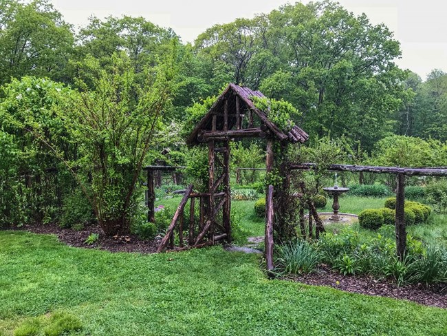 A wooden entryway into a fenced garden with a fountain in the middle.