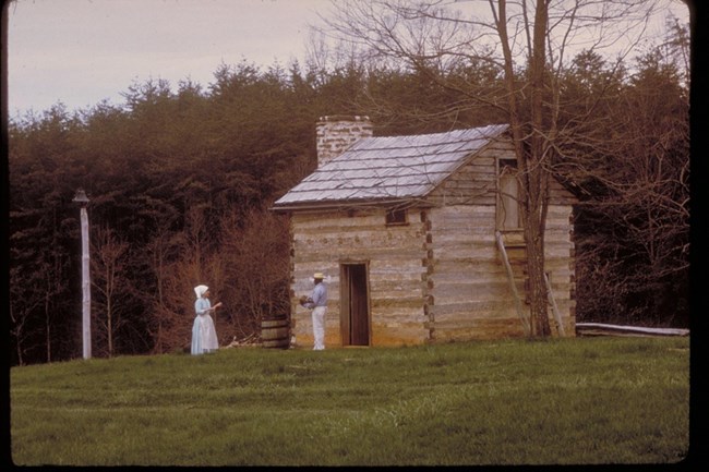 A small wooden cabin against a dense forest, with two people in period clothing standing in front.