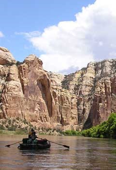 lt= An inflatable raft carrying two people and gear floats down a wide, calm river toward a towering wall of reddish sandstone with white clouds and blue sky overhead.