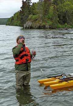 A researcher wearing green wading gear and an orange life vest stands knee-deep in water using a small device floating on small yellow pontoons. A researcher wearing green wading gear and an orange life vest stands knee-deep in water using a small device floating on small yellow pontoons.