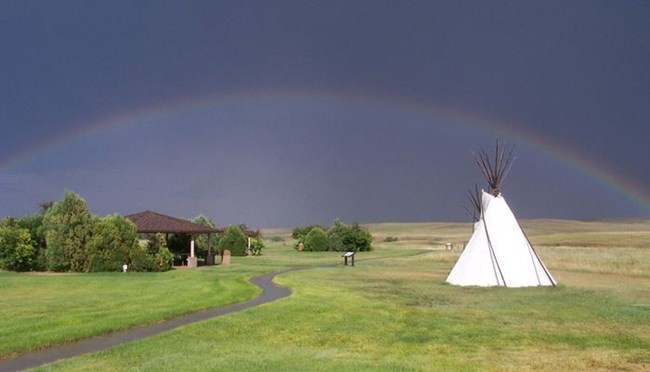 Rainbow and dark sky over green plains and white teepee.