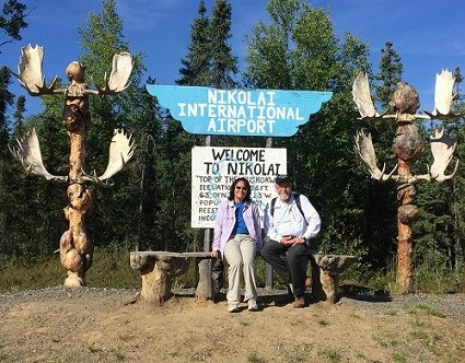 A man and a woman sitting on a rustic bench in front of a sign that says Nikolai International Airport. A man and a woman sitting on a rustic bench in front of a sign that says Nikolai International Airport.