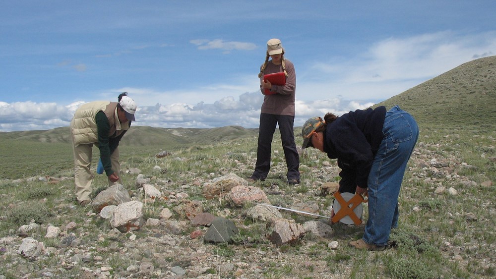 three people bent over a rocky area with grassy hills, blue sky, and clouds in the background three people bent over a rocky area with grassy hills, blue sky, and clouds in the background