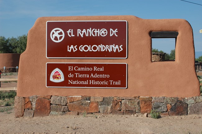 lt= A large brown sign at the entrance to el Golondrinas, wrapped in adobe.