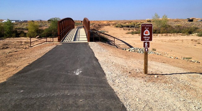 A paved trail leads to a bridge with brown railing. A brown sign stands on the right side of the pavement with a trail logo on it.