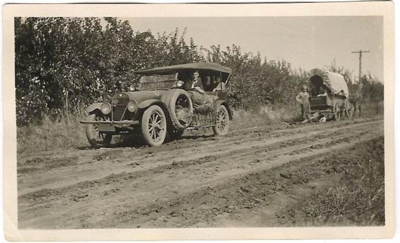 A brown toned photo of an old care on a road with a covered wagon behind it.