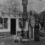 A historic image of a man standing next to an old gas station.