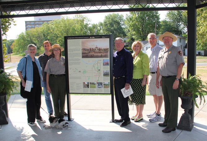 Seven people stand in front of a tall exhibit showing images and a map, surrounded by trees. Seven people stand in front of a tall exhibit showing images and a map, surrounded by trees.