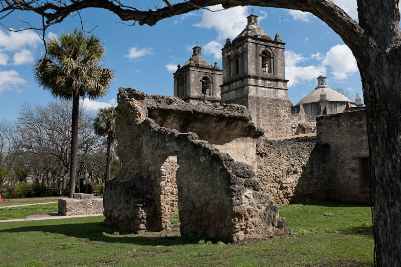 Old, weathered, stone building consisting of two fortress-like towers and a crumbling rock wall, which is surrounded by a grass lawn with palm trees.