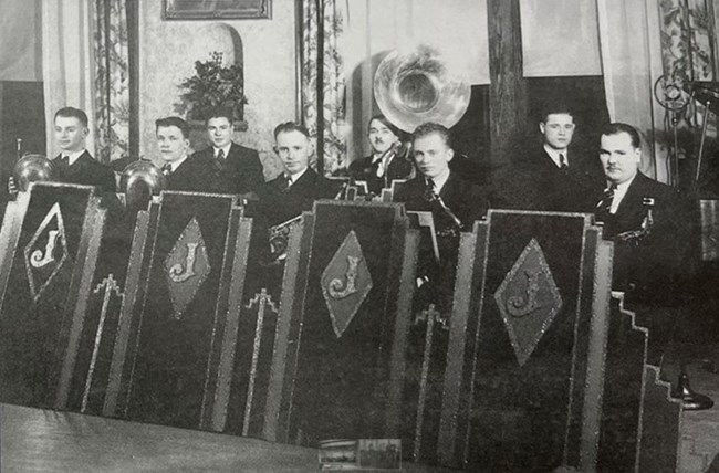 lt= A black and whit image of men in suits standing behind chairs.