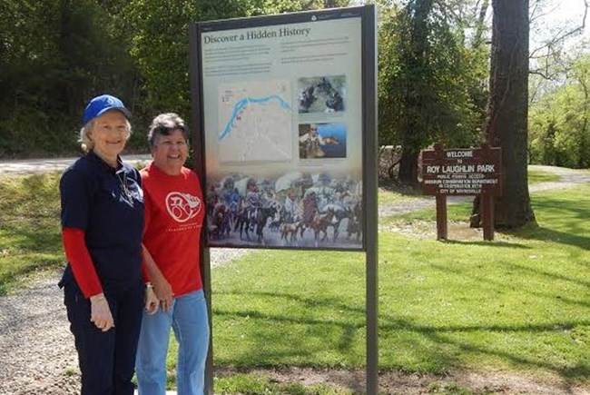 Two smiling women stand in front of an upright exhibit and in front of a green lawn with trees.