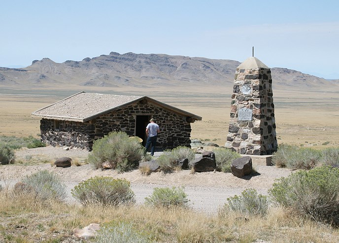 a house made of rock and a large rock monument in a desert landscape with mountains and blue sky in the background a house made of rock and a large rock monument in a desert landscape with mountains and blue sky in the background