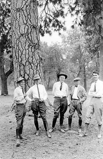 Five men in hats, ties, riding pants, and high boots stand under a tall evergreen tree.