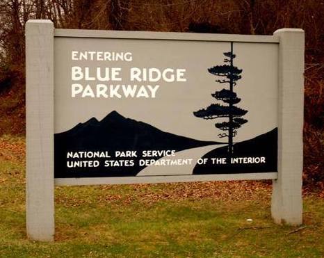 A wooden sign with a mountain, roadway, and tree marks an entrance to the Blue Ridge Parkway. NPS photo.