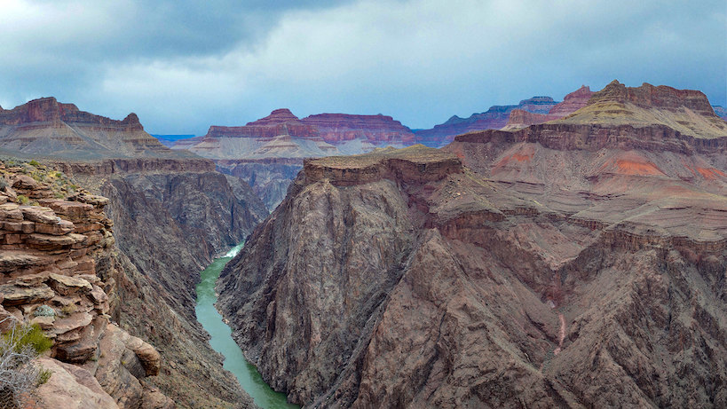 Colorado River as seen from Plateau Point off the Bright Angel Trail - Grand Canyon National Park. A green river flows beneath towering desert cliffs beneath an overcast sky.