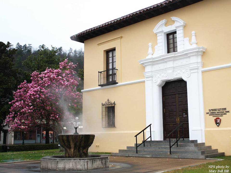 Hot Springs National Park in early springtime. NPS photo by M Hill. Steam rises from a water feature in front of a two-story building with a blossoming tree nearby.